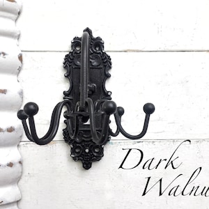 May include: Black metal wall hook with ornate detailing and four hooks. The hook is mounted on a white wall with the text "Dark Walnut" written below it.