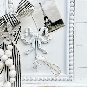 May include: White wooden board with a black and white striped ribbon, a white ceramic bow hook, a white beaded garland, a vintage postcard of the Eiffel Tower, and gold-rimmed glasses.