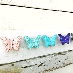 K&ouml;nnte beinhalten: Sechs schmetterlingsf&ouml;rmige Schrankkn&ouml;pfe in Pastellt&ouml;nen von Rosa, T&uuml;rkis, Teal, Lila und Grau. Sie sind auf einer wei&szlig;en, rustikalen Holzoberfl&auml;che montiert.