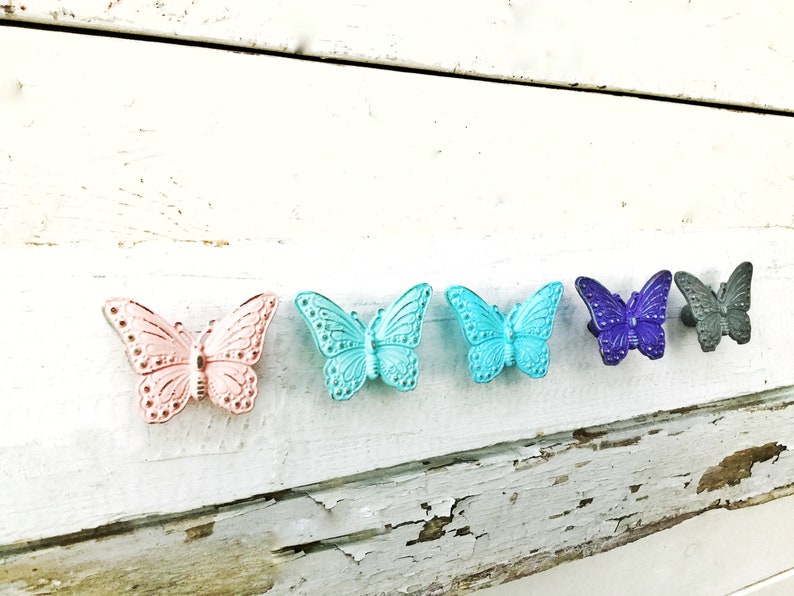 K&ouml;nnte beinhalten: Sechs schmetterlingsf&ouml;rmige Schrankkn&ouml;pfe in Pastellt&ouml;nen von Rosa, T&uuml;rkis, Teal, Lila und Grau. Sie sind auf einer wei&szlig;en, rustikalen Holzoberfl&auml;che montiert.