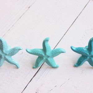 May include: Three turquoise blue starfish ornaments on a white wooden background. The starfish are different shades of blue, with the one on the left being the lightest and the one on the right being the darkest.