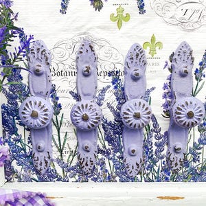 May include: Four lavender-colored metal wall hooks with a distressed finish. Each hook features a decorative floral knob and a detailed backplate. The hooks are set against a backdrop of lavender sprigs and botanical prints.