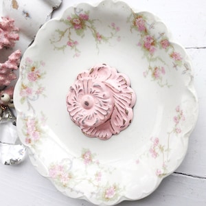 May include: A pink floral decorative knob with a white center, sitting on a white porcelain plate with a floral pattern.