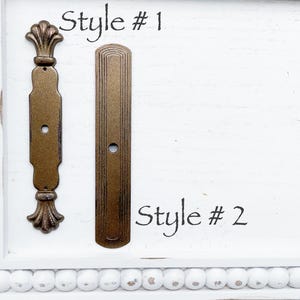 May include: Two antique bronze-colored cabinet pulls, labeled "Style #1" and "Style #2", are displayed on a white, distressed wooden surface. Style #1 features ornate detailing, while Style #2 has a simple, rectangular design.