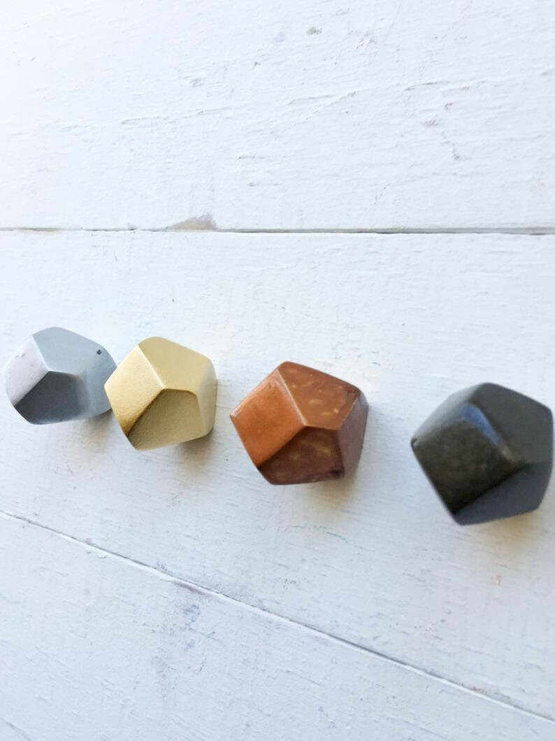 May include: Four geometric shaped ceramic knobs in gray, gold, copper, and black. The knobs are mounted on a white wooden surface.