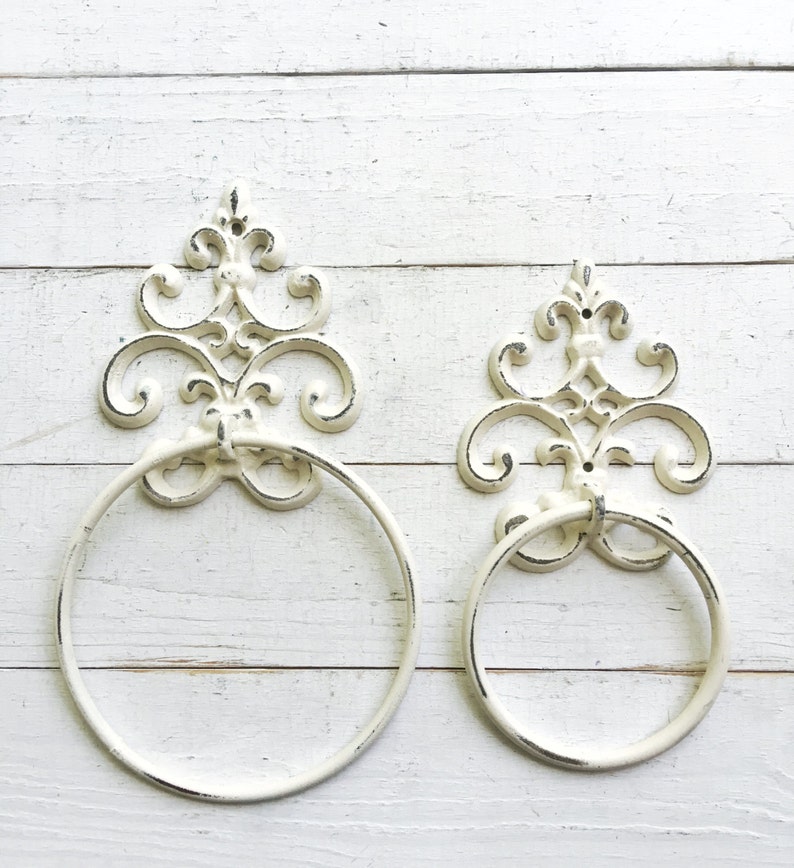 Peut inclure: Deux anneaux de serviette en m&eacute;tal blanc avec des motifs de volutes orn&eacute;s. Les anneaux sont fix&eacute;s &agrave; un mur en bois blanc.