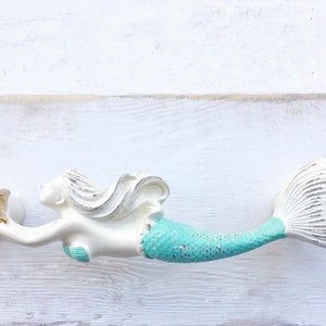 May include: White mermaid figurine with a turquoise tail and a gold starfish in its hand. The mermaid is holding the starfish up in the air. The figurine is on a white wooden background.