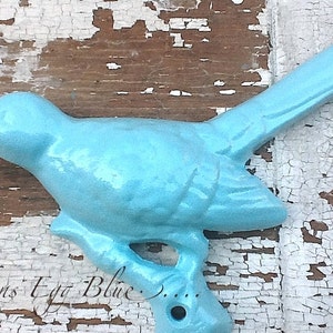 Puede incluir: Una figurita de pájaro azul claro de color azul huevo de petirrojo. La figurita es de metal y tiene un aspecto rústico y vintage. El pájaro está posado en una rama con un agujero en la parte inferior. El texto "Robins Egg Blue" está escrito en la parte inferior de la imagen.