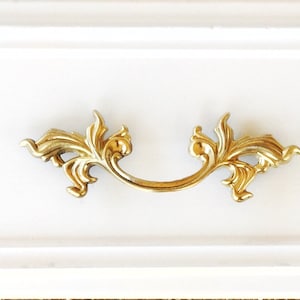 May include: A white drawer with a gold ornate pull handle. The handle has a decorative design with a leaf motif.