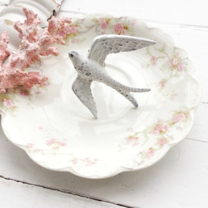 May include: A white porcelain plate with a scalloped edge and a delicate floral pattern in pink and green. A white metal bird figurine with outstretched wings sits in the center of the plate.