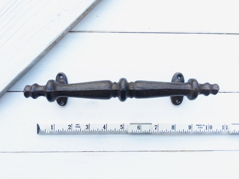 May include: A dark brown, cast iron door pull with a decorative design. The door pull is approximately 10 inches long and has mounting holes on each end. The hardware is shown on a white background with a ruler for scale.