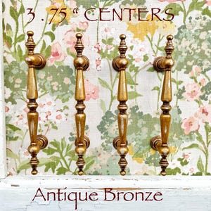 May include: Four antique bronze cabinet pulls with a 3.75-inch center-to-center measurement. The pulls have a decorative design with a gold finish and white accents. The background features a floral pattern.