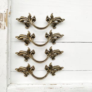 May include: Four ornate gold-toned drawer pulls with a decorative scroll design. The pulls are arranged in a vertical row on a white wooden surface.