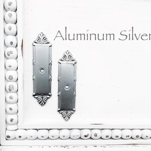 May include: Two aluminum silver door plates with ornate detailing are displayed on a white surface. The text "Aluminum Silver" is above the plates. The door plates have a rectangular shape with a circular indentation in the center.