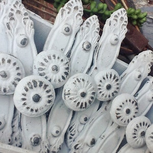 K&ouml;nnte beinhalten: Eine Sammlung von wei&szlig;en, verzierten, metallenen Schubladengriffen mit einer abgenutzten Oberfl&auml;che. Jeder Griff hat ein dekoratives Design und einen runden Knopf mit einer erhabenen Mitte.