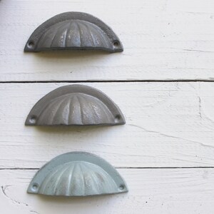 May include: Three cabinet pulls in different shades of gray. The pulls are made of metal and have a scalloped design.