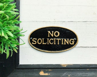 May include: A black oval sign with gold lettering that reads "NO SOLICITING" on a white wooden background.