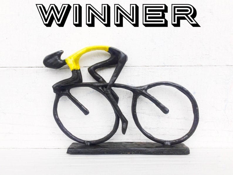 May include: Black metal sculpture of a cyclist in a yellow jersey riding a bicycle. The sculpture is on a black base and the word "WINNER" is written above the sculpture.