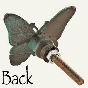 K&ouml;nnte beinhalten: Dekorativer Schrankknopf in Schmetterlingsform mit einem Used-Look-Finish. Der Knopf hat einen bronzefarbenen K&ouml;rper mit gr&uuml;nen Akzenten auf den Fl&uuml;geln. Das Wort "Back" ist in schwarzer Schrift geschrieben. Die Gewindeschraube erstreckt sich von hinten.