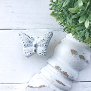 May include: A white painted butterfly-shaped drawer pull with a distressed finish. The pull is mounted on a white painted surface with a distressed finish.