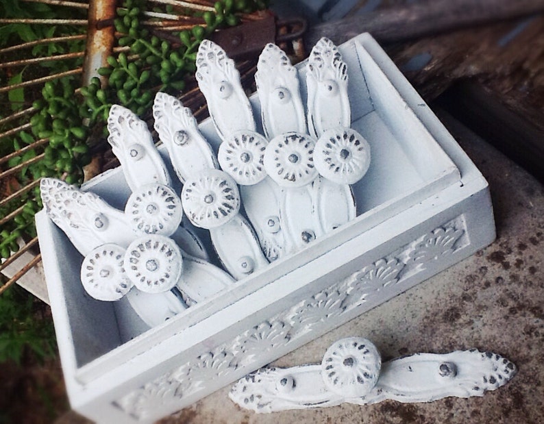 K&ouml;nnte beinhalten: Eine wei&szlig;e Holzkiste mit einem dekorativen Blumendesign, gef&uuml;llt mit wei&szlig;en Metall-Schubladengriffen mit silbernen Akzenten. Die Schubladengriffe haben einen Vintage-Stil und sind in einer Reihe angeordnet.