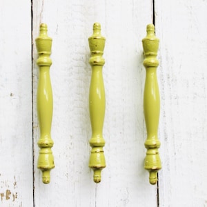 May include: Three yellow wooden drawer pulls with a decorative top. The pulls are on a white wooden background.