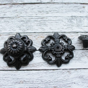 May include: Three black metal decorative knobs with ornate designs. The knobs have a floral pattern and are suitable for furniture or cabinetry.
