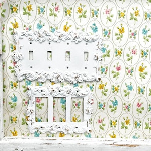 May include: Two white porcelain light switch plates with decorative floral trim. The plates are mounted on a white wall with a floral wallpaper pattern.