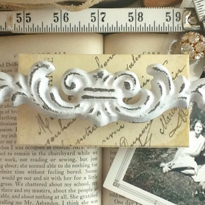 May include: A white metal decorative drawer pull with a scroll design. The pull is on a piece of paper with handwritten text.