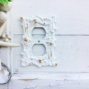 May include: White porcelain outlet cover with ornate gold accents. The cover has a decorative design with a floral motif.