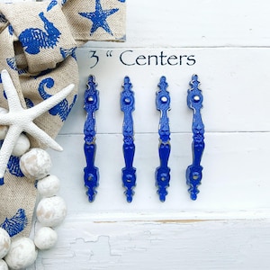 May include: Four cobalt blue cabinet pulls with a decorative design, arranged vertically on a white wooden surface. The text "3" Centers" is visible. A nautical-themed bow and starfish are in the background.