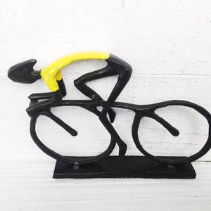 May include: Black metal sculpture of a cyclist in a yellow jersey riding a bicycle. The sculpture is on a black base.