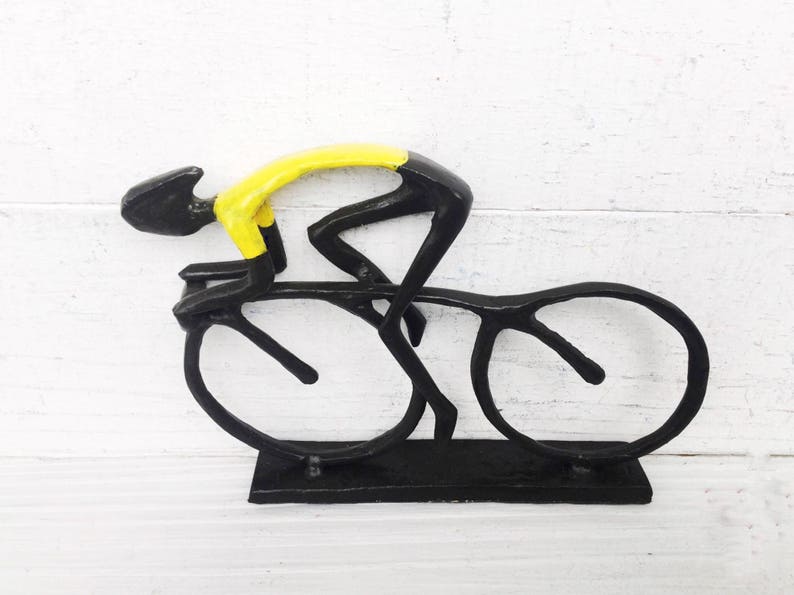 May include: Black metal sculpture of a cyclist in a yellow jersey riding a bicycle. The sculpture is on a black base.