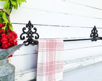 May include: Black wrought iron towel rack with decorative scrollwork on the ends. A red and white checkered kitchen towel is hanging on the rack.