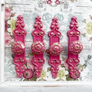 May include: Four pink door knobs with ornate gold detailing and a floral pattern. The knobs are displayed on a white surface with a floral pattern.
