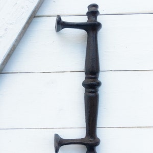 May include: A black cast iron door pull with a vertical handle and two horizontal arms. The handle has a decorative, ornate design. The door pull is set against a white wooden background.