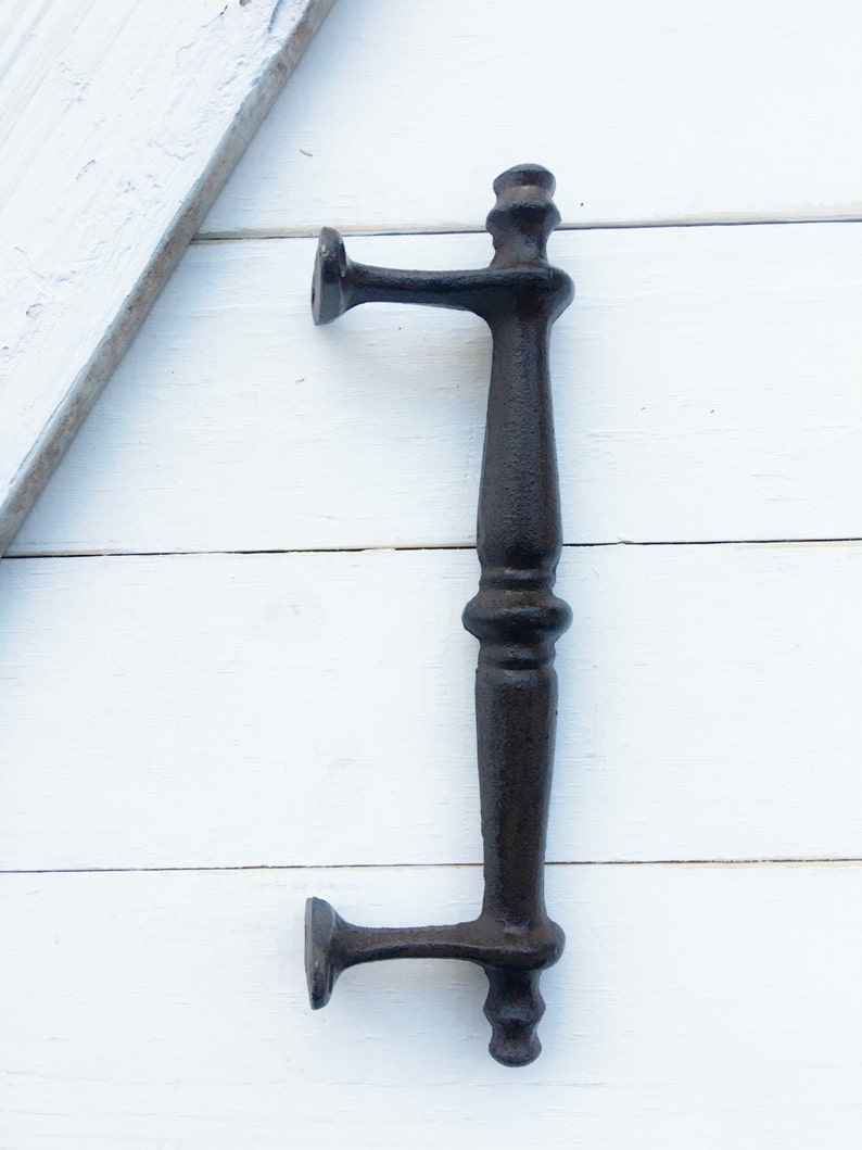 May include: A black cast iron door pull with a vertical handle and two horizontal arms. The handle has a decorative, ornate design. The door pull is set against a white wooden background.