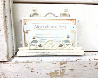 May include: White metal business card holder with a decorative scroll design. The holder has a card insert with the text "Alacartcreations" printed on it.