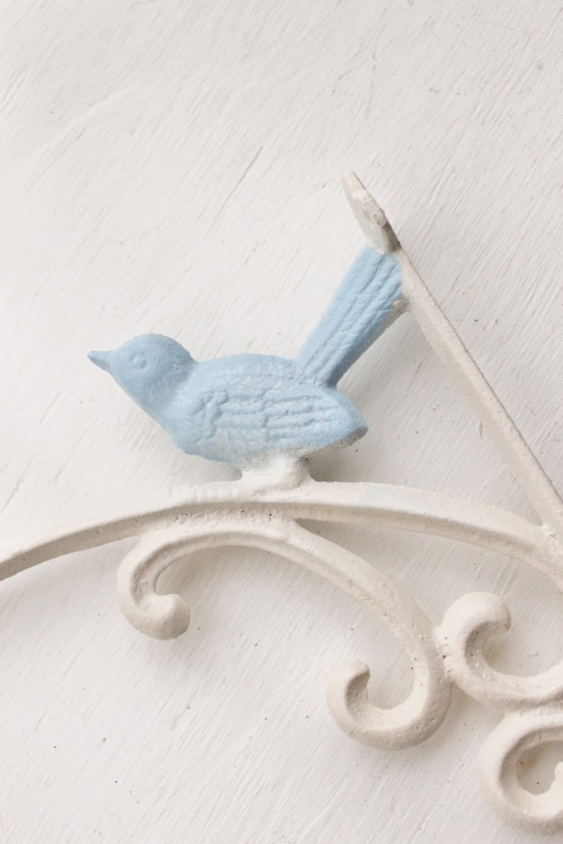 May include: White metal shelf bracket with a blue bird design. The bracket has a decorative scrollwork design.