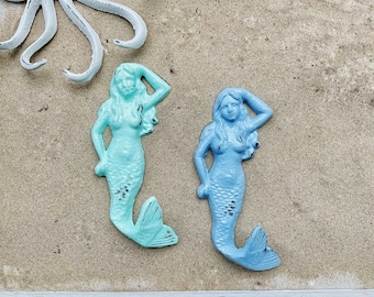 May include: Two mermaid-shaped decorative items in shades of blue and teal, with a distressed finish. The mermaids are posed with one arm raised. A white octopus-shaped item is in the background. The items are on a sandy surface.