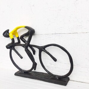 May include: Black metal sculpture of a cyclist in a yellow jersey riding a bicycle. The sculpture is on a white wooden surface.