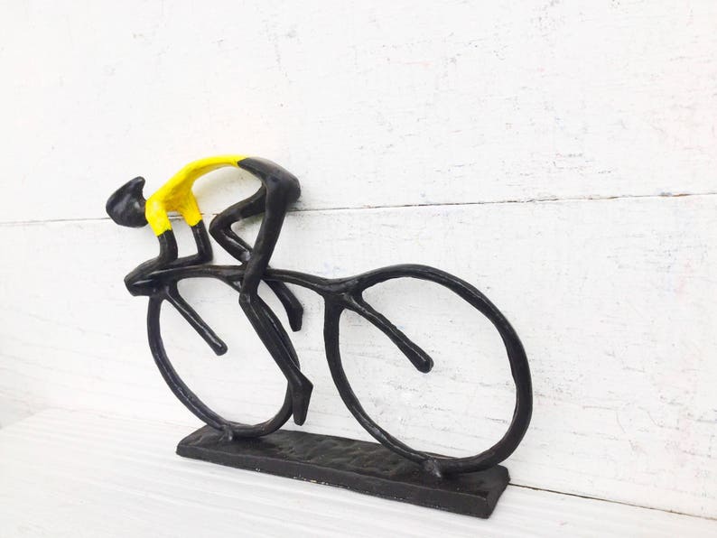 May include: Black metal sculpture of a cyclist in a yellow jersey riding a bicycle. The sculpture is on a white wooden surface.