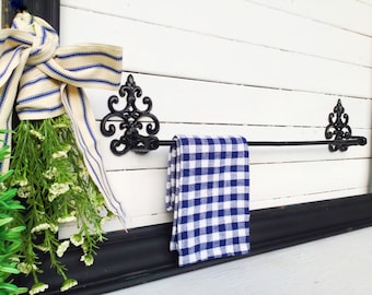 May include: Black wrought iron towel rack with decorative scrollwork on the ends. A blue and white checkered kitchen towel is hanging on the rack.