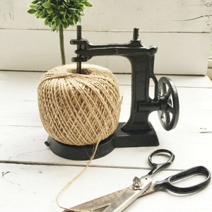 May include: A black cast iron twine dispenser with a ball of twine and a small green plant on top. A pair of silver scissors are lying on a white wooden surface.