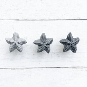May include: Three gray starfish on a white wooden background. The starfish are arranged in a row, with the points of the starfish facing the viewer.