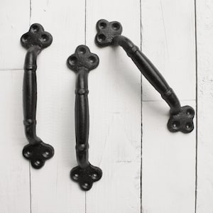May include: Three black metal door handles with a decorative, quatrefoil-shaped design. The handles are on a white wooden background.