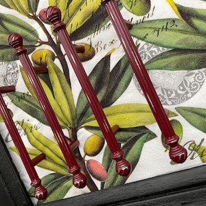 May include: A set of four burgundy-colored cabinet pulls with decorative finials. The pulls are arranged on a tray with a botanical print featuring olive branches and olives. The tray has a black frame.