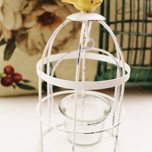 May include: White metal birdcage candle holder with a yellow bird perched on top. The candle holder has a glass votive holder inside.