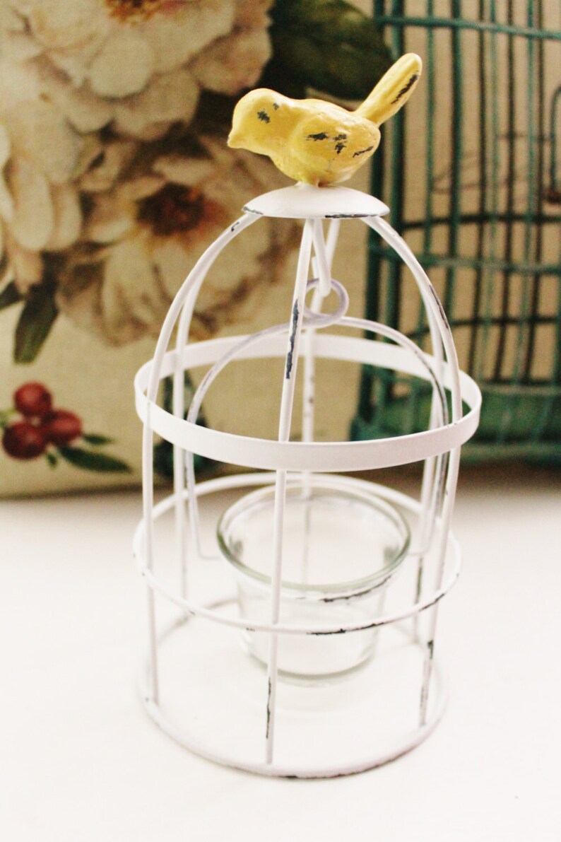 May include: White metal birdcage candle holder with a yellow bird perched on top. The candle holder has a glass votive holder inside.
