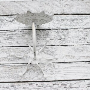 May include: White metal coat rack with a decorative top and five hooks. The rack is mounted on a white wooden wall.